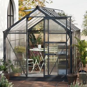 Greenhouse with glass panels and metal frame, surrounded by plants and outdoor furniture.